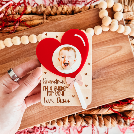 Wooden magnet with a heart design and a photo of a baby, held by a hand on a wooden surface.